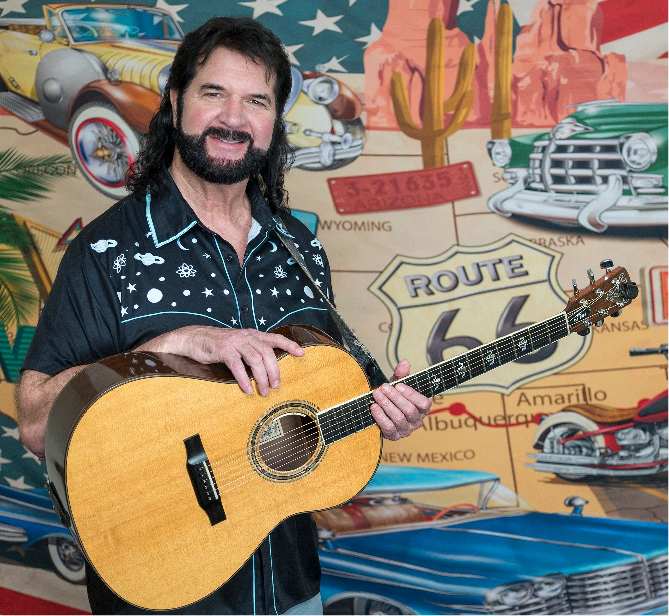 Man with beard and guitar in front of colorful Route 66 mural.