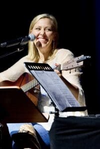 Smiling woman playing guitar and singing into a microphone.